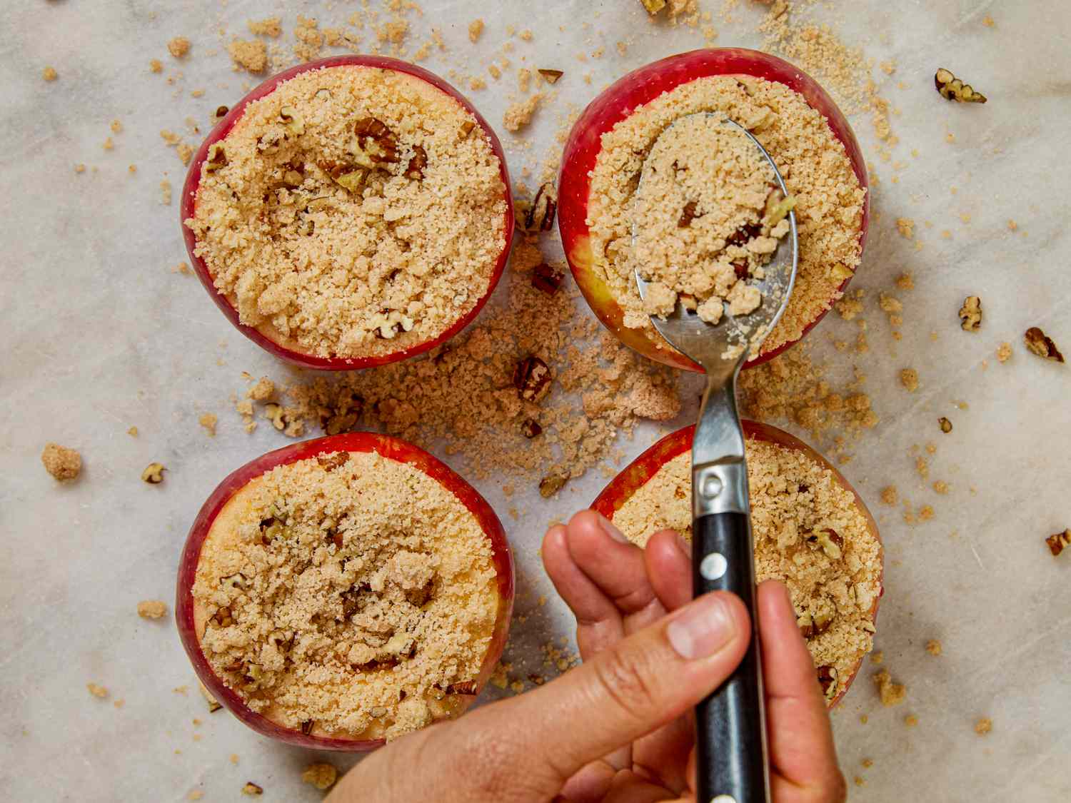 Four baked apples with a crumb topping a hand holding a spoon scooping some of the topping