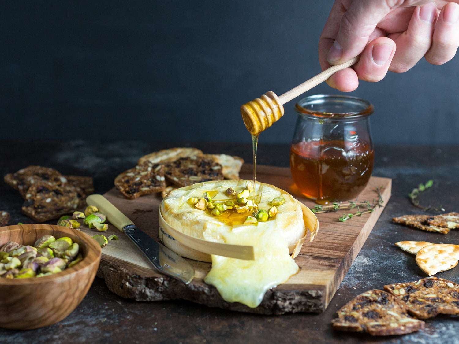 Author drizzling honey on a wheel of baked brie, topped with pistachio and served with crackers. 