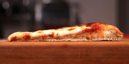 A cross-section of a New York-style pizza slice with under-developed dough, showing flat texture and rise.