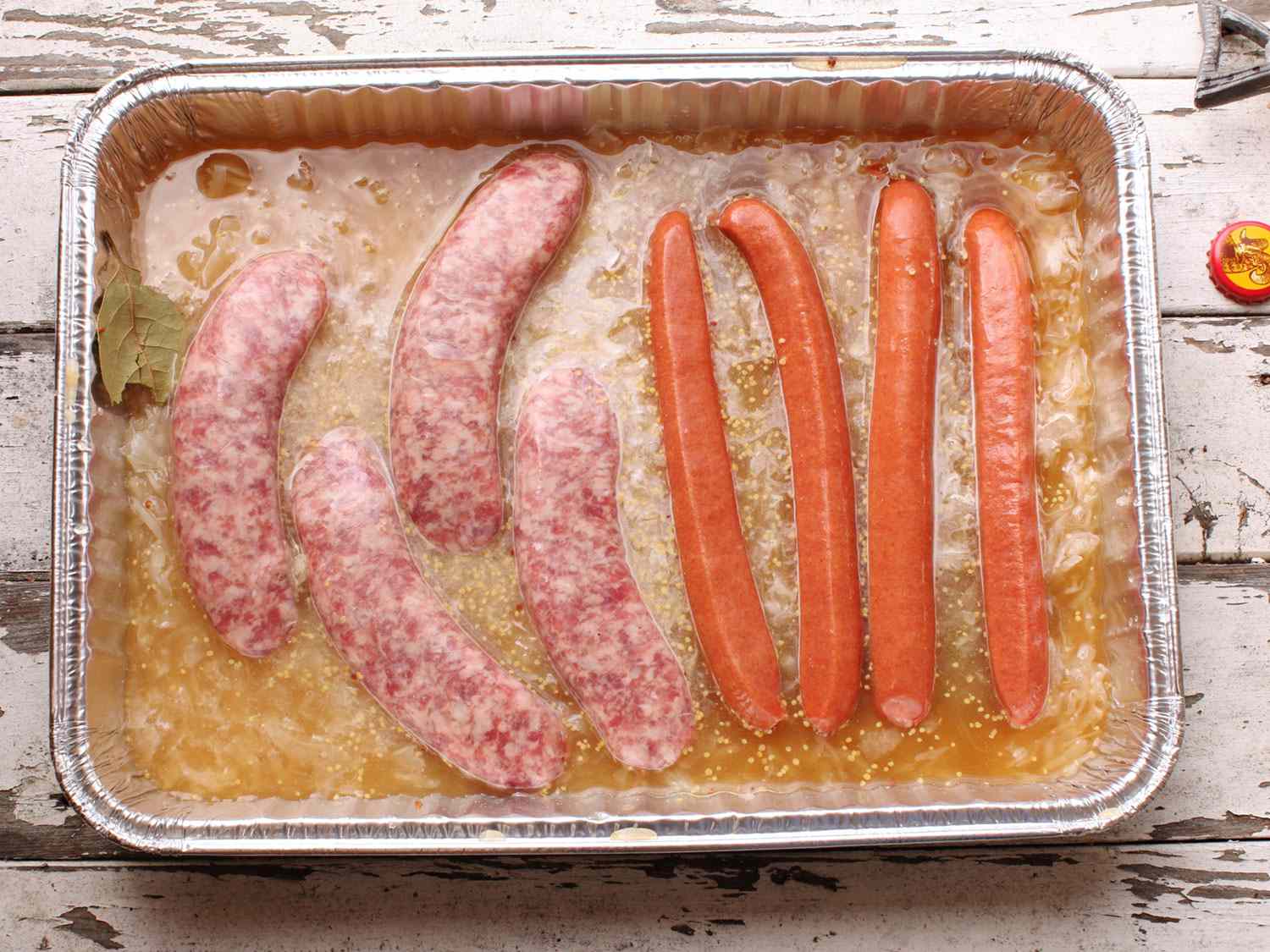 Overhead view of sausages and hot dogs partially submerged in a disposable aluminum pan filled with a flavorful braising liquid.