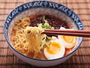 A bowl of prepared ramen from Sun Noodles. The ramen has a two soft-boiled egg halves, green onions, and broth. A pair of chopsticks are lifting some of the noodles. 