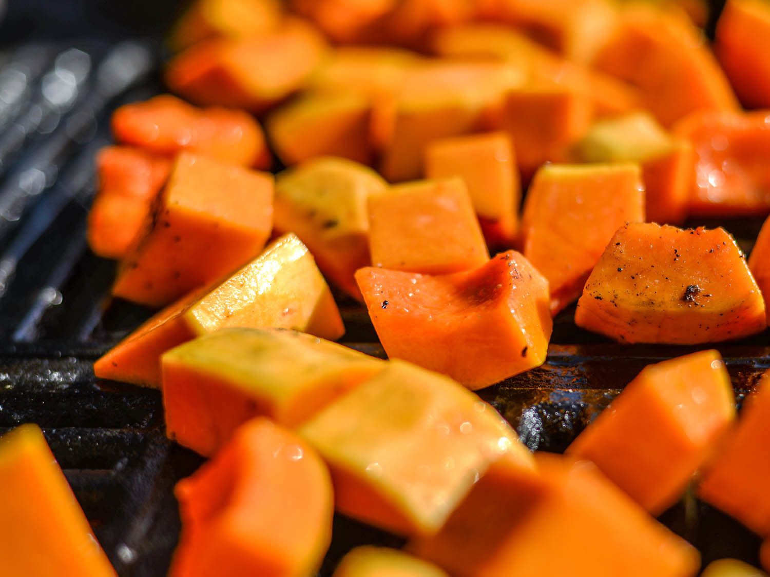 Butternut squash cubes glistening with oil and pepper on a grill grate. 