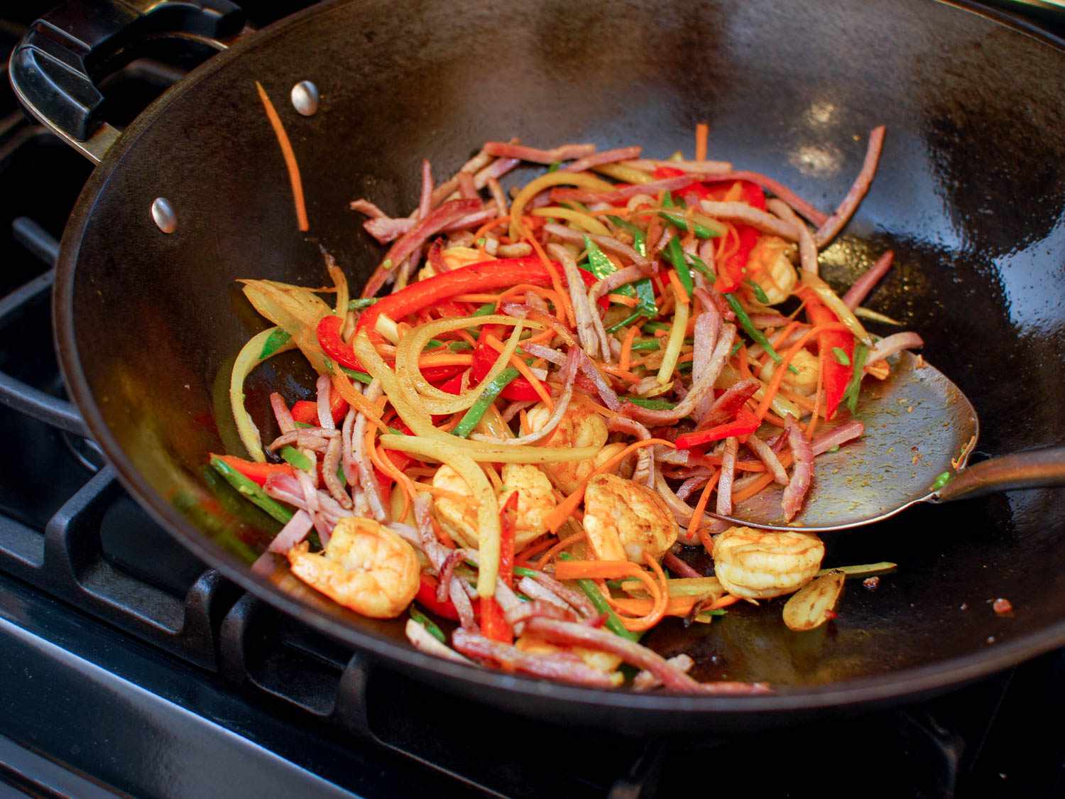 A metal spatula stirring a wok full of shrimp, pork, onion, bell peppers, snow peas, carrots, and curry powder.