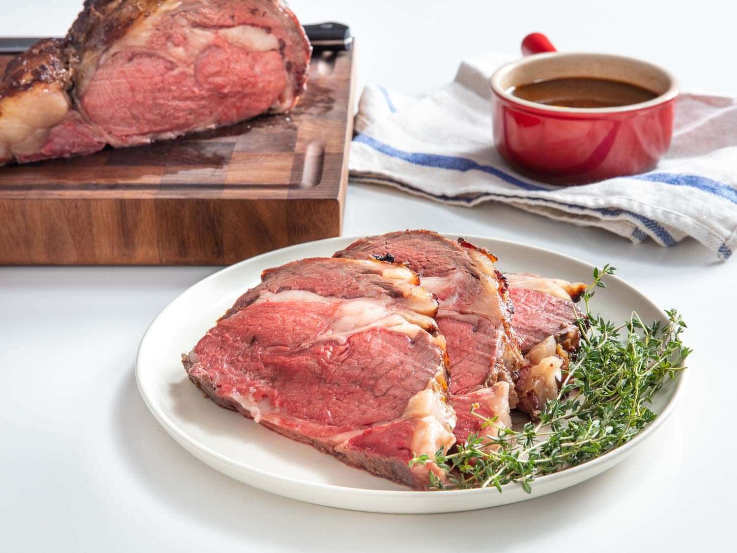 Side view closeup of three thick slices of koji prime rib on a serving plate with thyme sprigs, and the remaining prime rib on a cutting board in the background along with a bowl of jus.