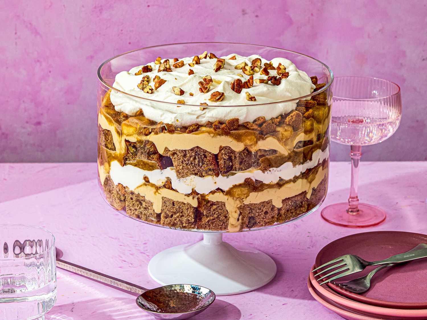 Layered banana bread trifle in a glass bowl garnished with whipped cream and nuts served with utensils and tableware on a pink surface