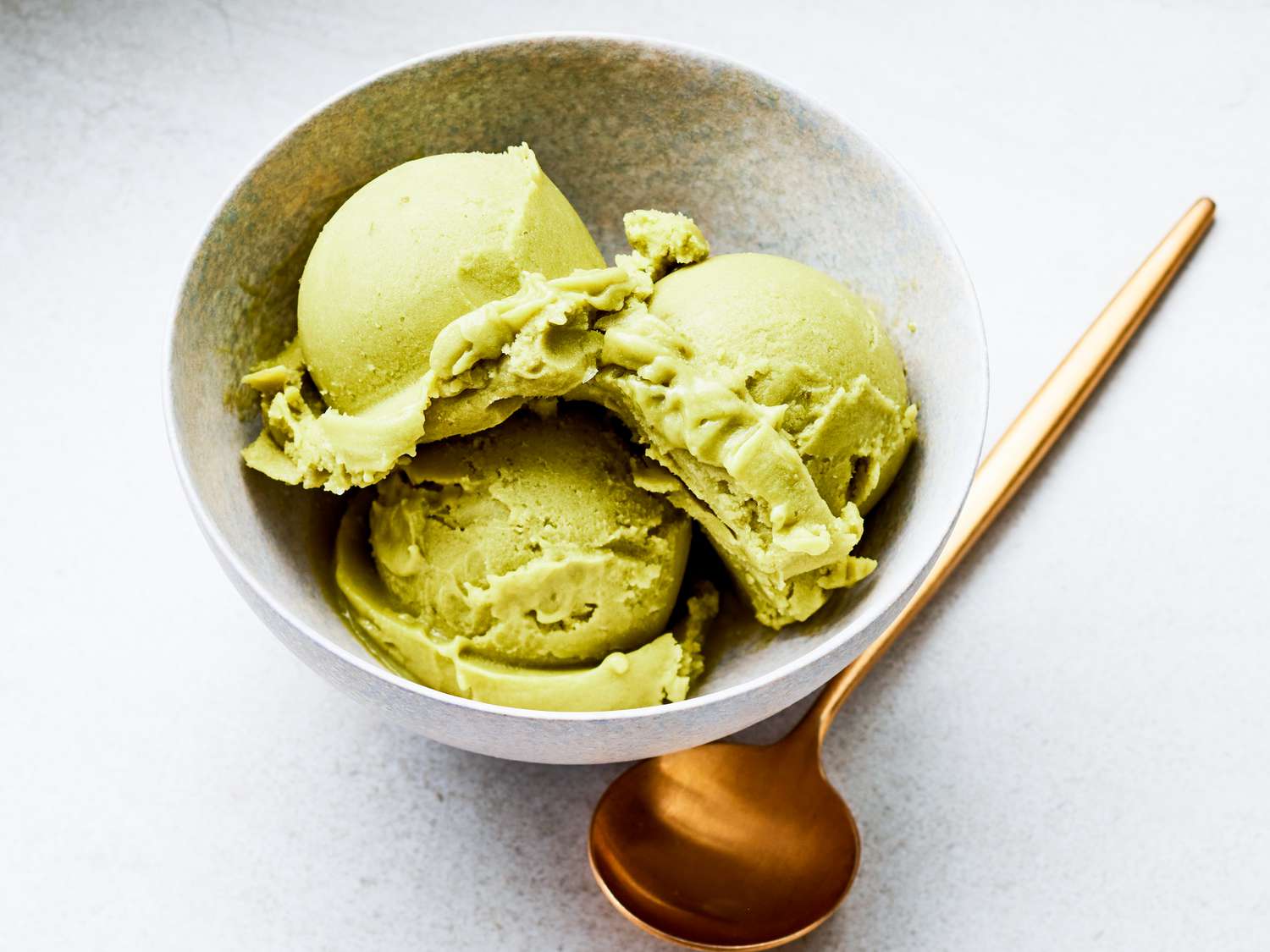 Three scoops of avocado ice cream in with bowl and gold spoon on the side