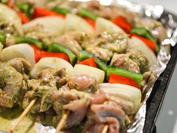 Raw marinated chicken, green and red bell pepper squares, and onion threaded onto skewers and placed on an aluminum foil-lined sheet tray.