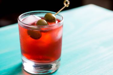 A Campari Spritz in a rocks glass, garnished with 3 olives on a pick. 