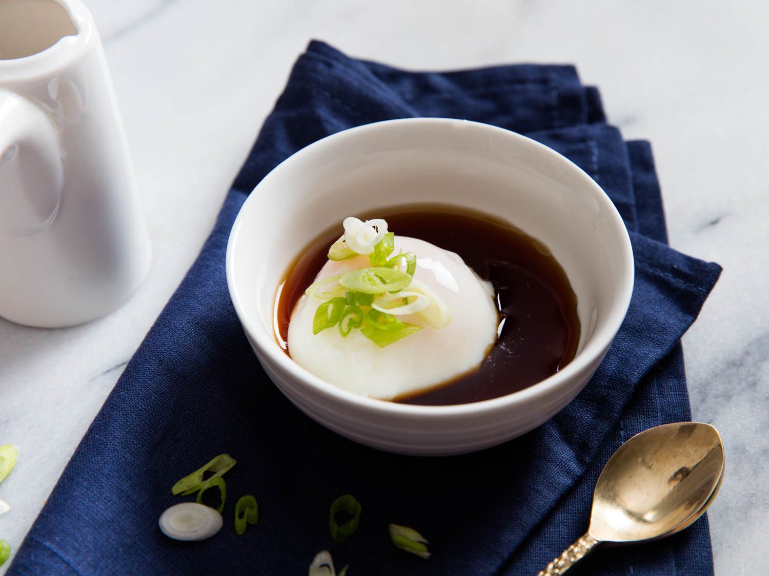 An onsen egg served at the table, cradled in a white bowl, half-submerged in soy-seasoned dashi. Sliced scallion is sprinkled over the top.