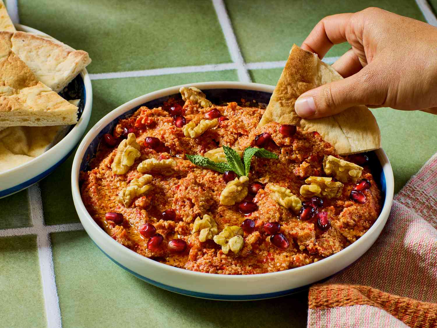 A dish of muhammara garnished with walnuts and herbs, someone dipping bread into the spread