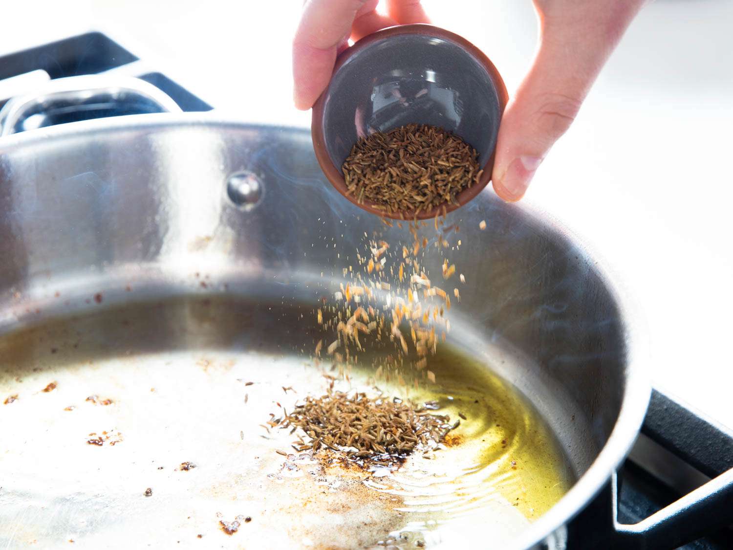 A hand adding cumin seeds from a small pinch bowl into a sauté pan