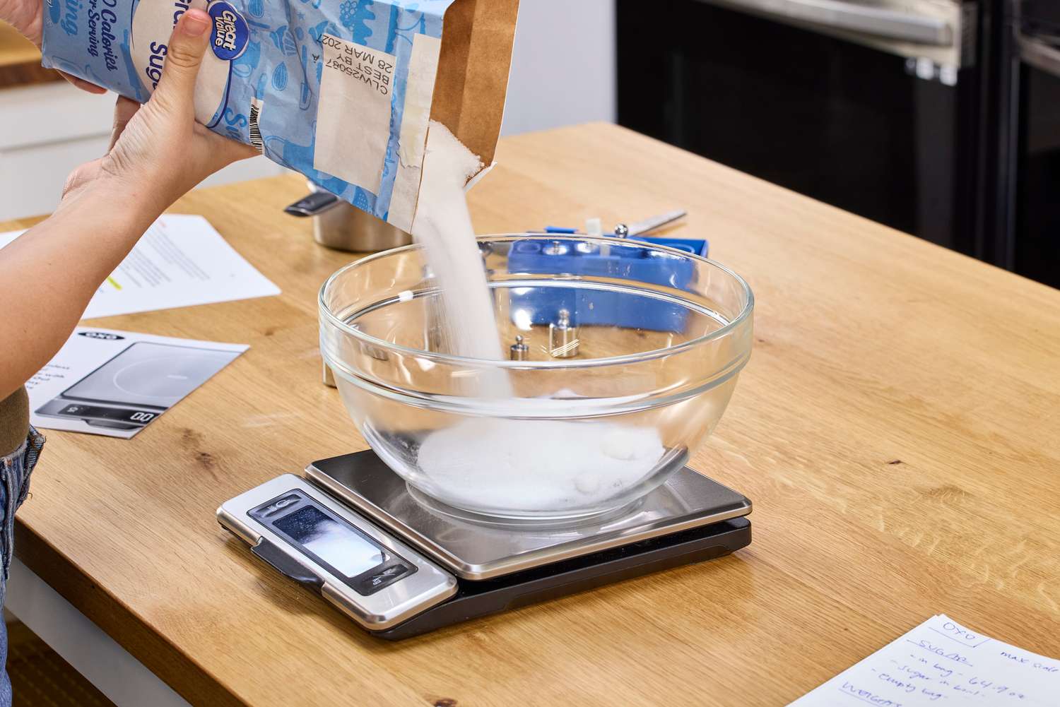 A person pours white sugar into a glass bowl on the OXO Good Grips Stainless Steel Food Scale with Pull-Out Display