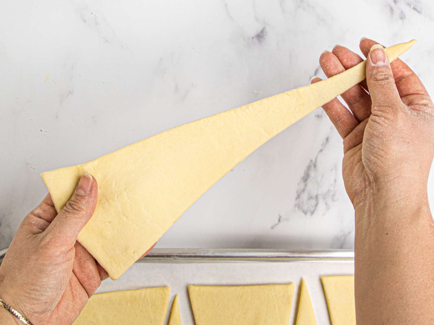 Hands folding a croissant dough triangle for shaping tray with dough triangles in the background