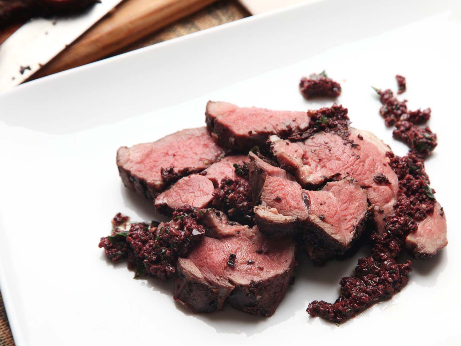 Closeup of sliced sous vide leg of lamb with black olives served on a white plate.