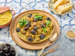 Lamb and prune Tagine with side of couscous, prunes, bread and cutlery on a blue tile surface 