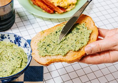A hand spreading green butter on a slice of toast with a knife a dish of butter bacon and scrambled eggs in the background