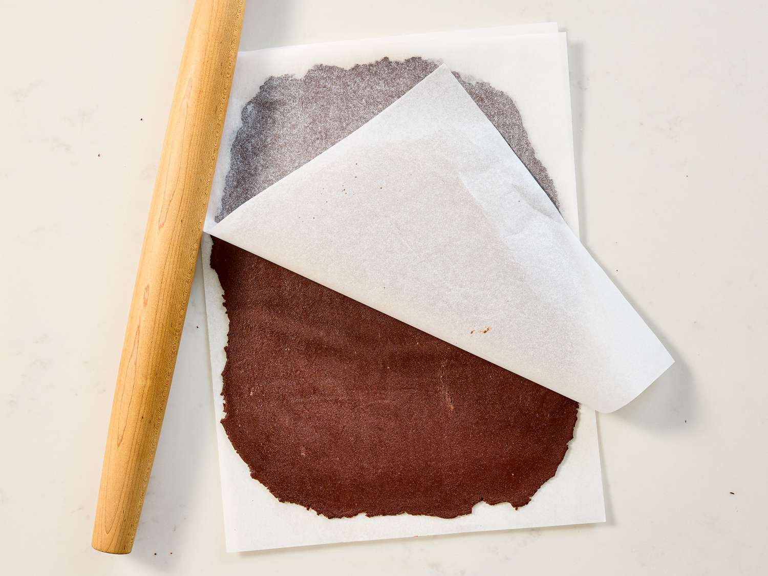 Flat rolled chocolate dough between parchment paper next to a wood rolling pin