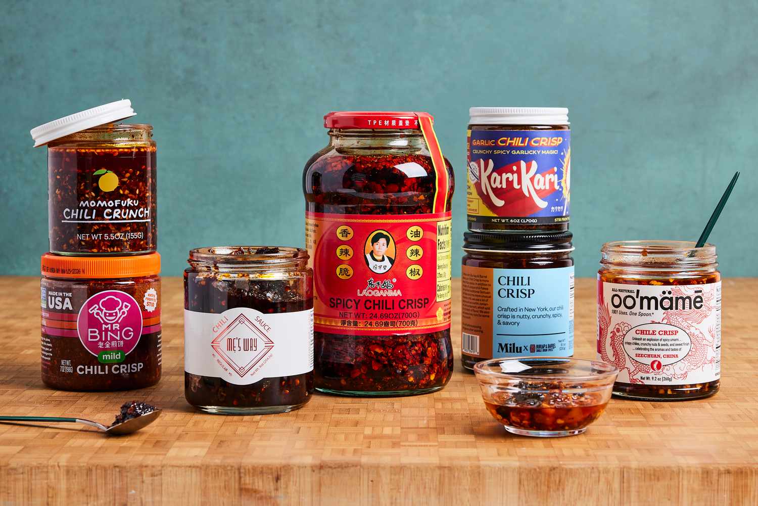 Numerous bottles of chili crisp on a wooden countertop. Some have their lids off.