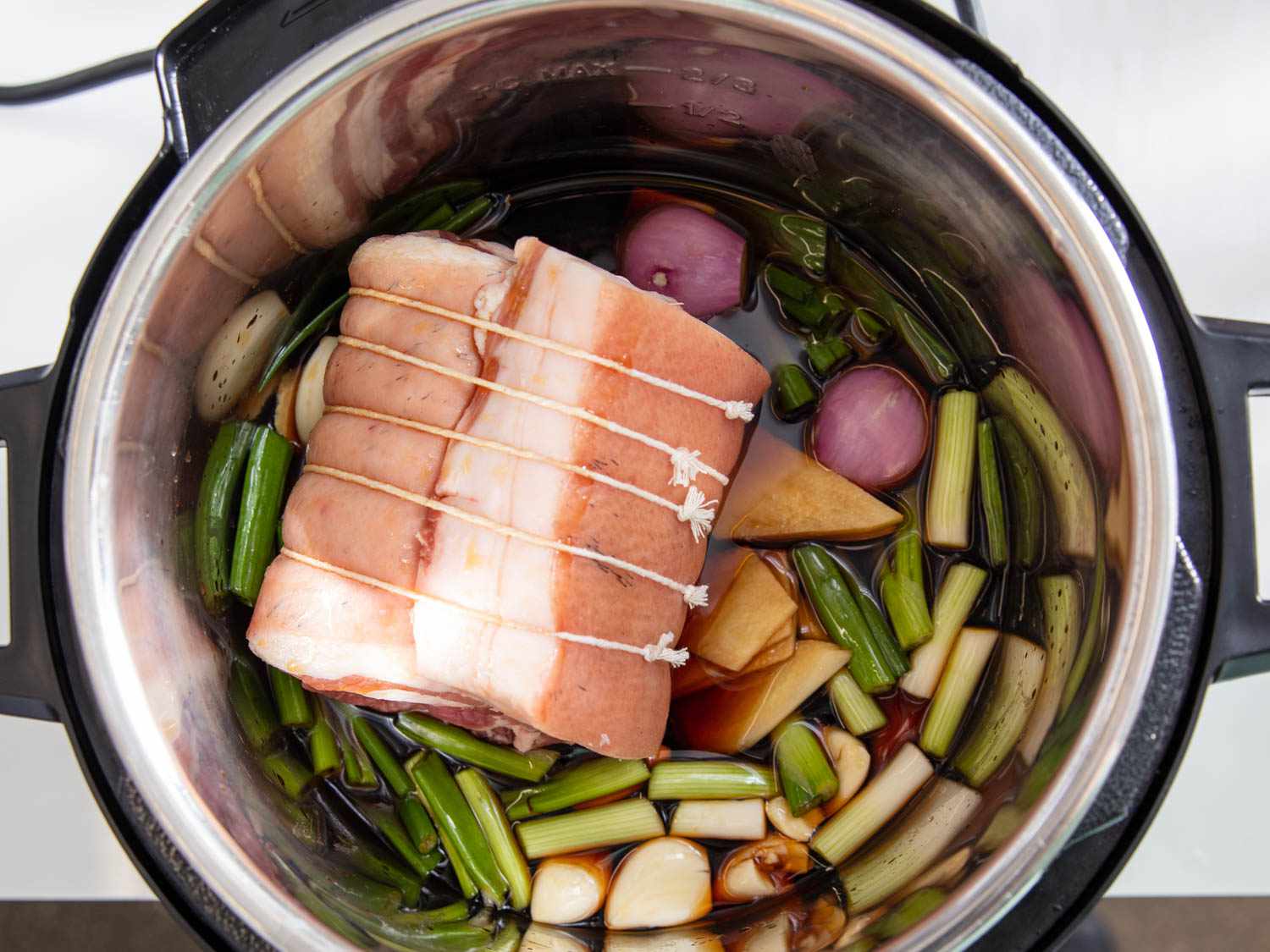 Overhead view of tied up roll of skin-on pork belly nestled on top of aromatics in a pressure cooker.