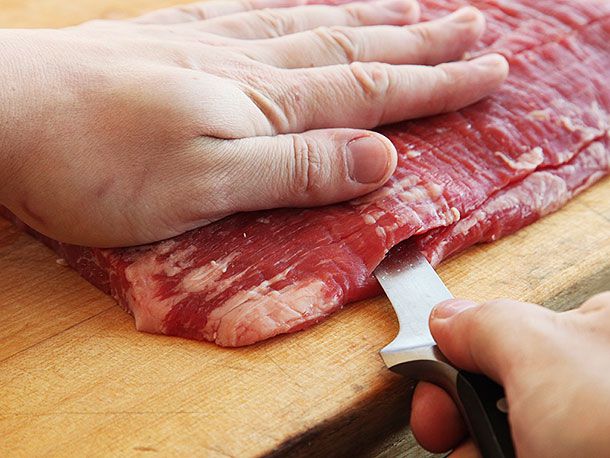 Hand pressing down on piece of raw flank steak as it's cut in half horizontally