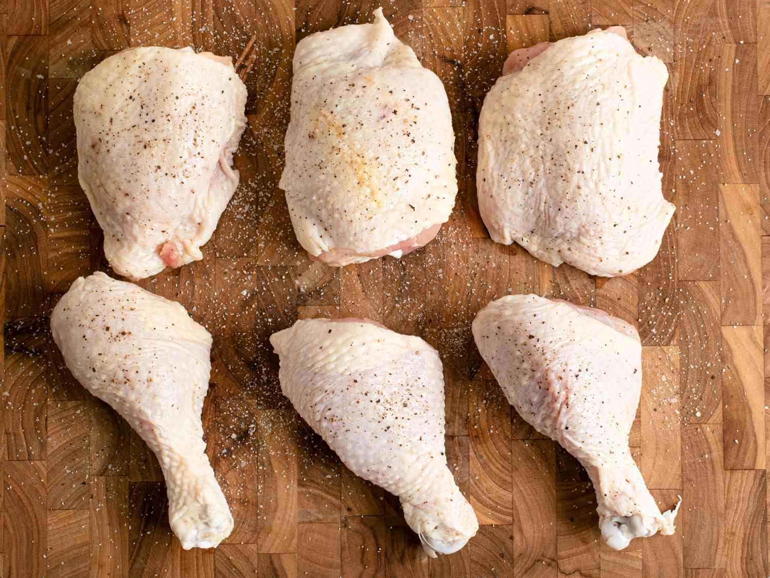 Three raw, skin-on, bone-in chicken thighs and three raw, skin-on, bone-in chicken legs placed on a wooden cutting board. The chicken is seasoned with salt and pepper.