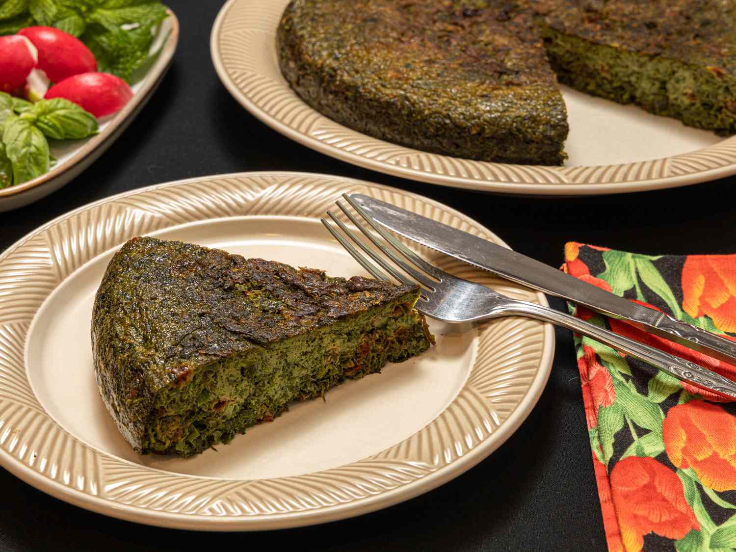 Serving of Herb Kookoo on a beige plate with utensils and a colorful napkin. Larger serving of Herb Kookoo in background 