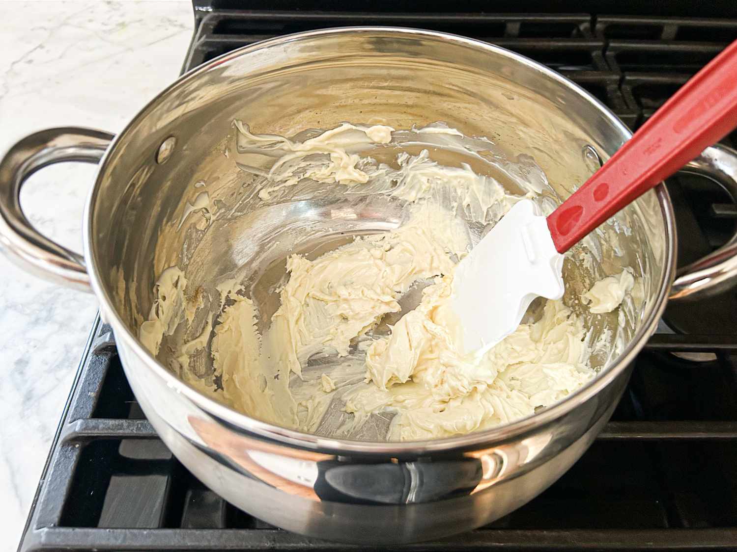 A pot on a stove containing melted white chocolate a redhandled spatula rests inside