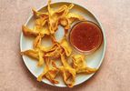 Golden, fried crab rangoons on a blue ceramic plate which also holds a small glass bowl of sauce, on a pink stone background.