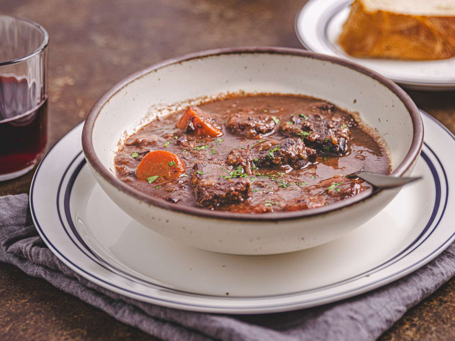 Bowl of stew on a plate on top of a blue napkin, with a glass of red wine, and a piece of bread on the background 