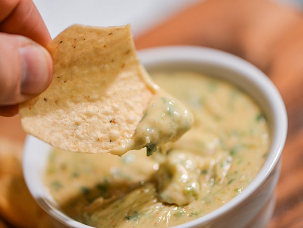 A tortilla chip dipped in green chile queso is held up to the camera.