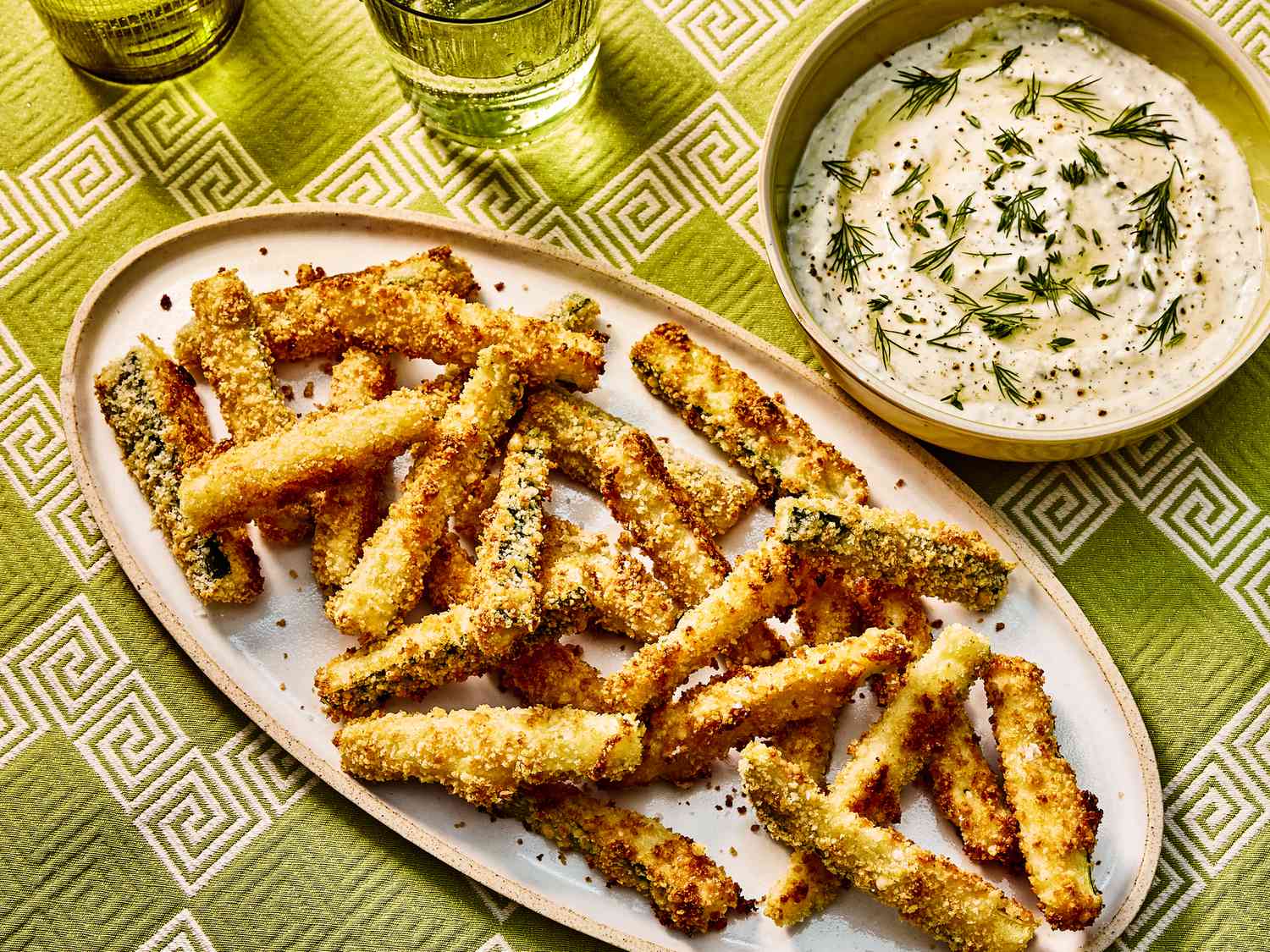 Baked Zucchini fries on a white dish, with a bowl of dip on the side. The table has green printed table cloth and two glasses of water. 