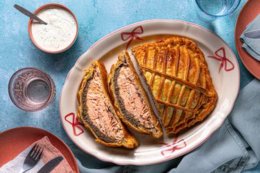 Salmon encased in pastry on a serving dish with dill sauce and tableware on a blue surface