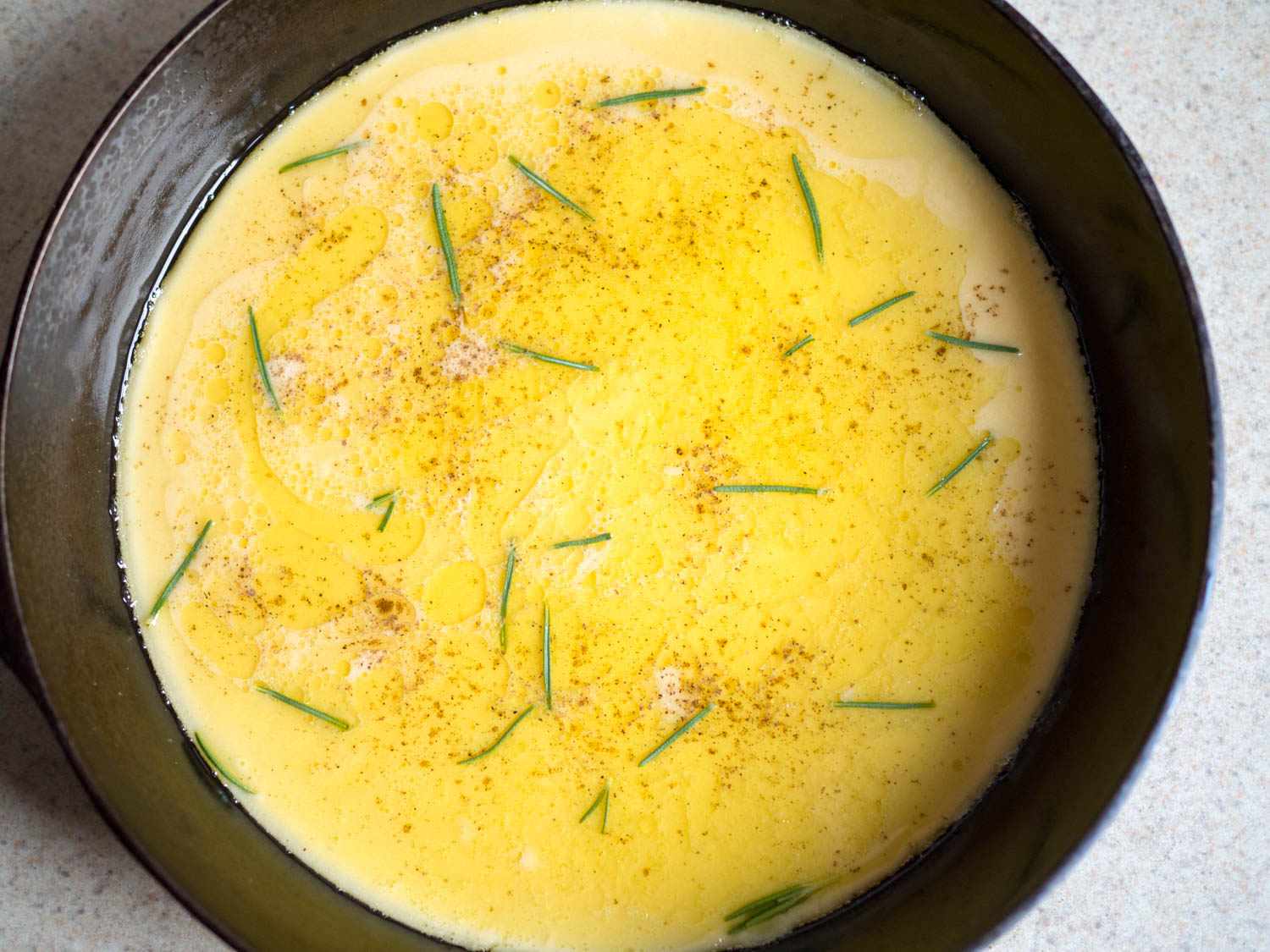 Farinata batter in a cast iron skillet with specks of pepper and rosemany leaves. 