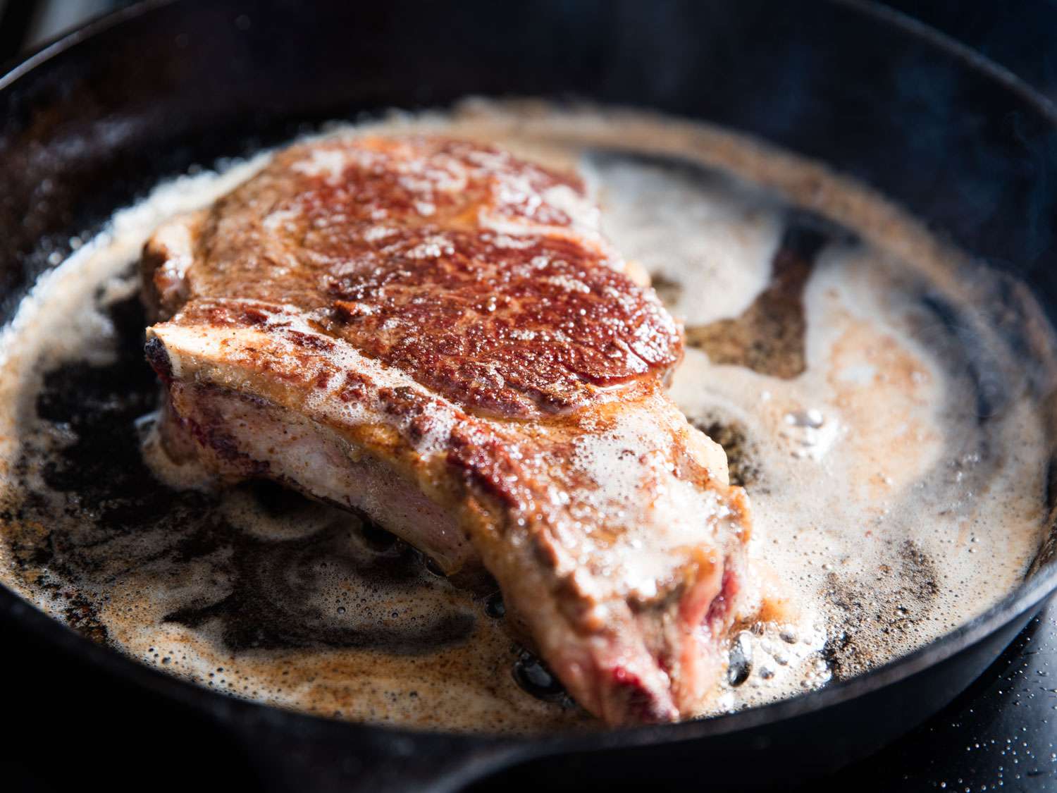 A bone in steak cooking in a cast iron skillet. 