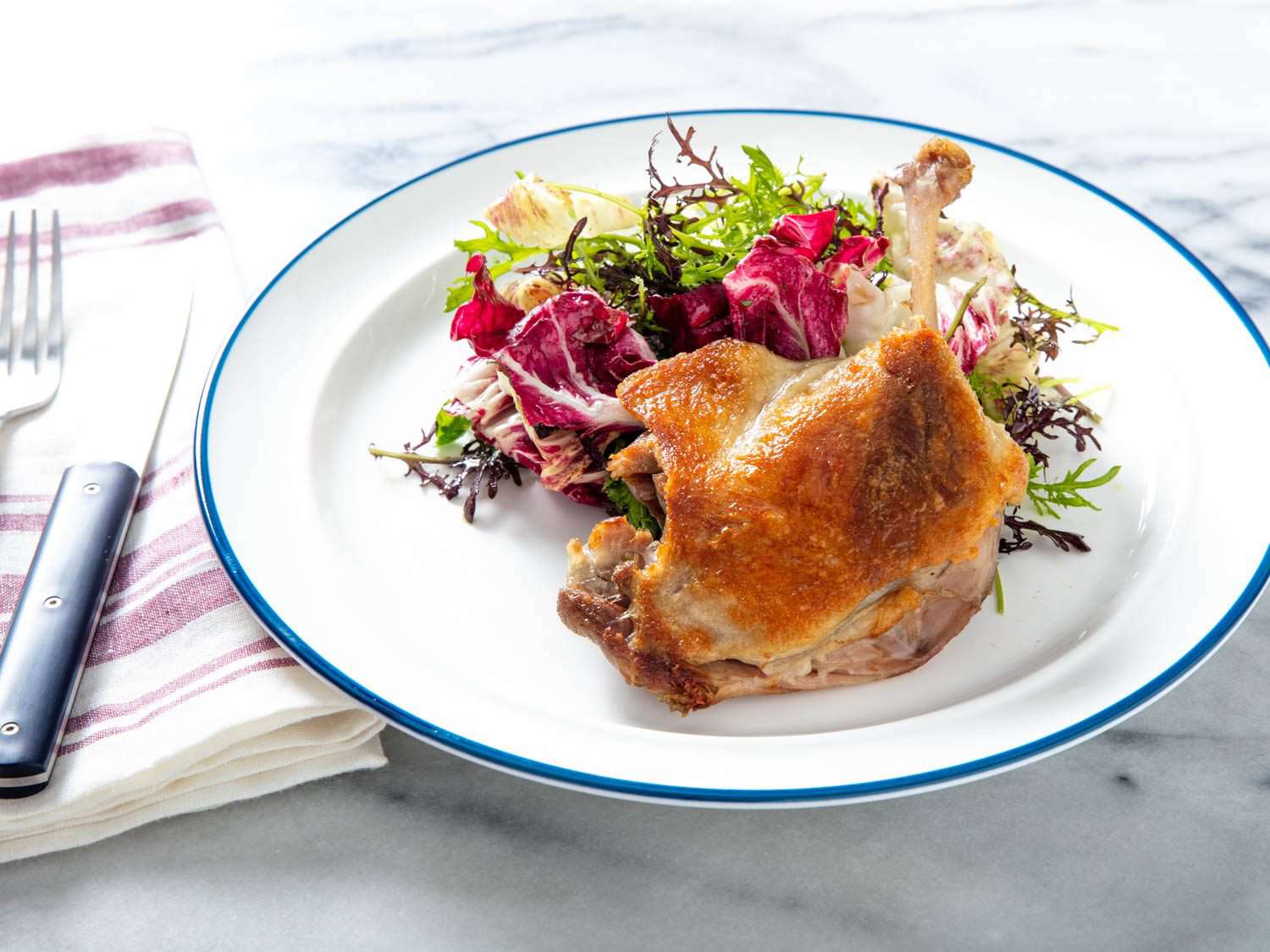 Closeup front view of a crisped confit duck leg with a mixed greens salad.