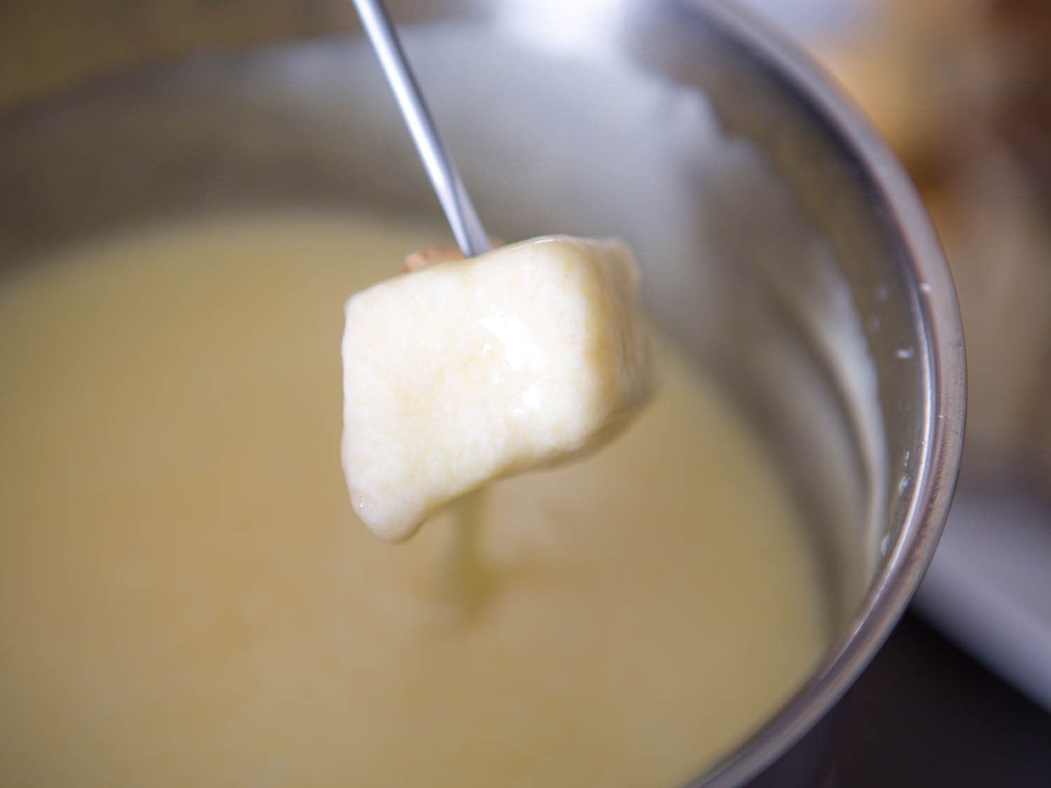 A cube of toast dipped in cheese fondue, resting above the pot.