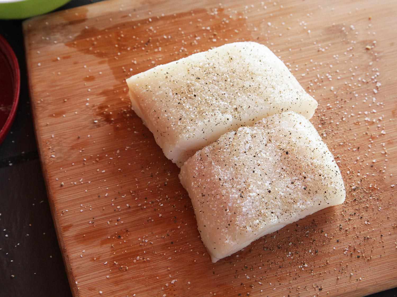 Two portion-sized pieces of white-fleshed fish fillet, arranged on a cutting board and showered with salt and pepper. 