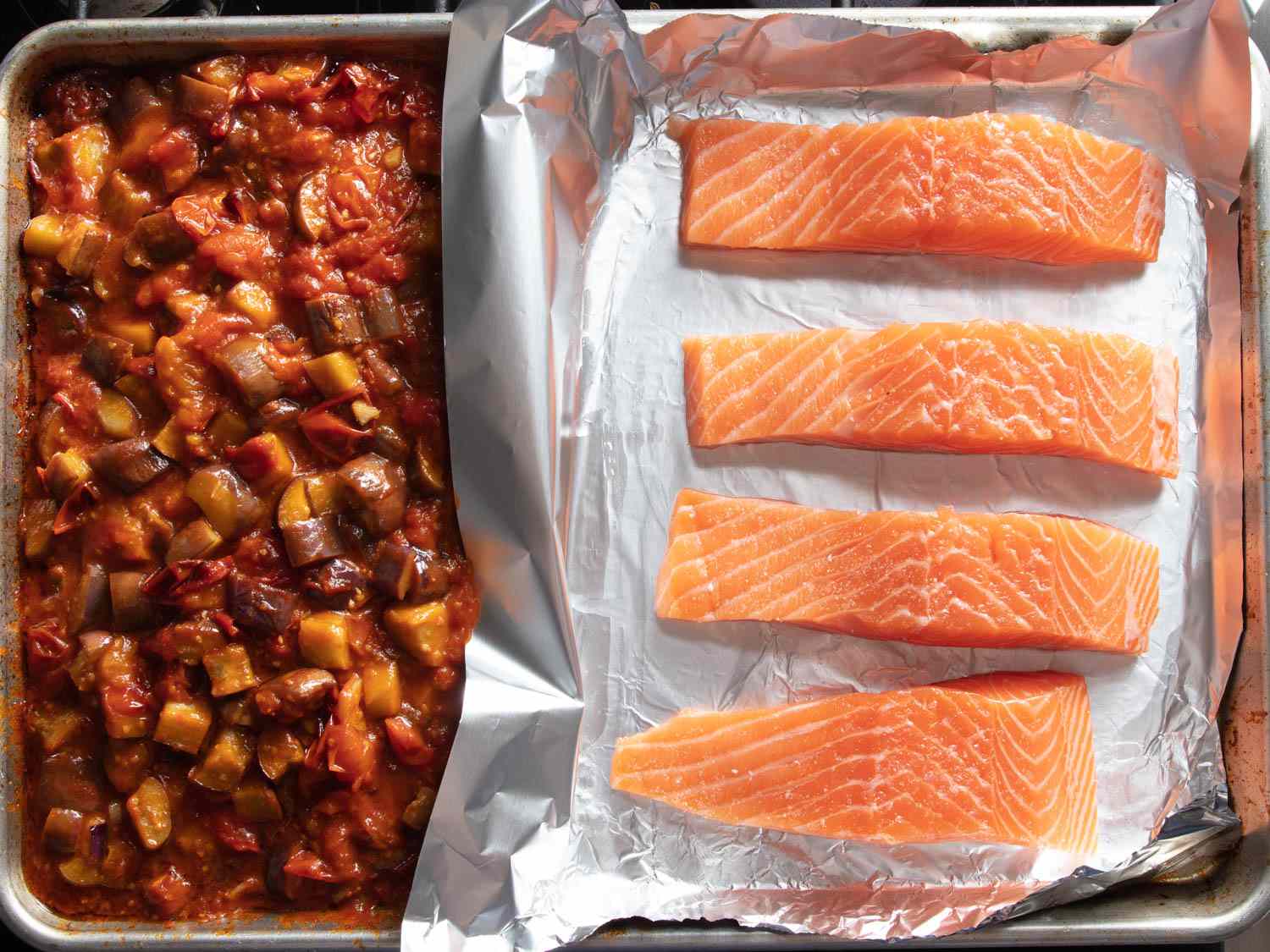 Chunks of raw salmon on foil next to tomato eggplant stew on a sheet pan