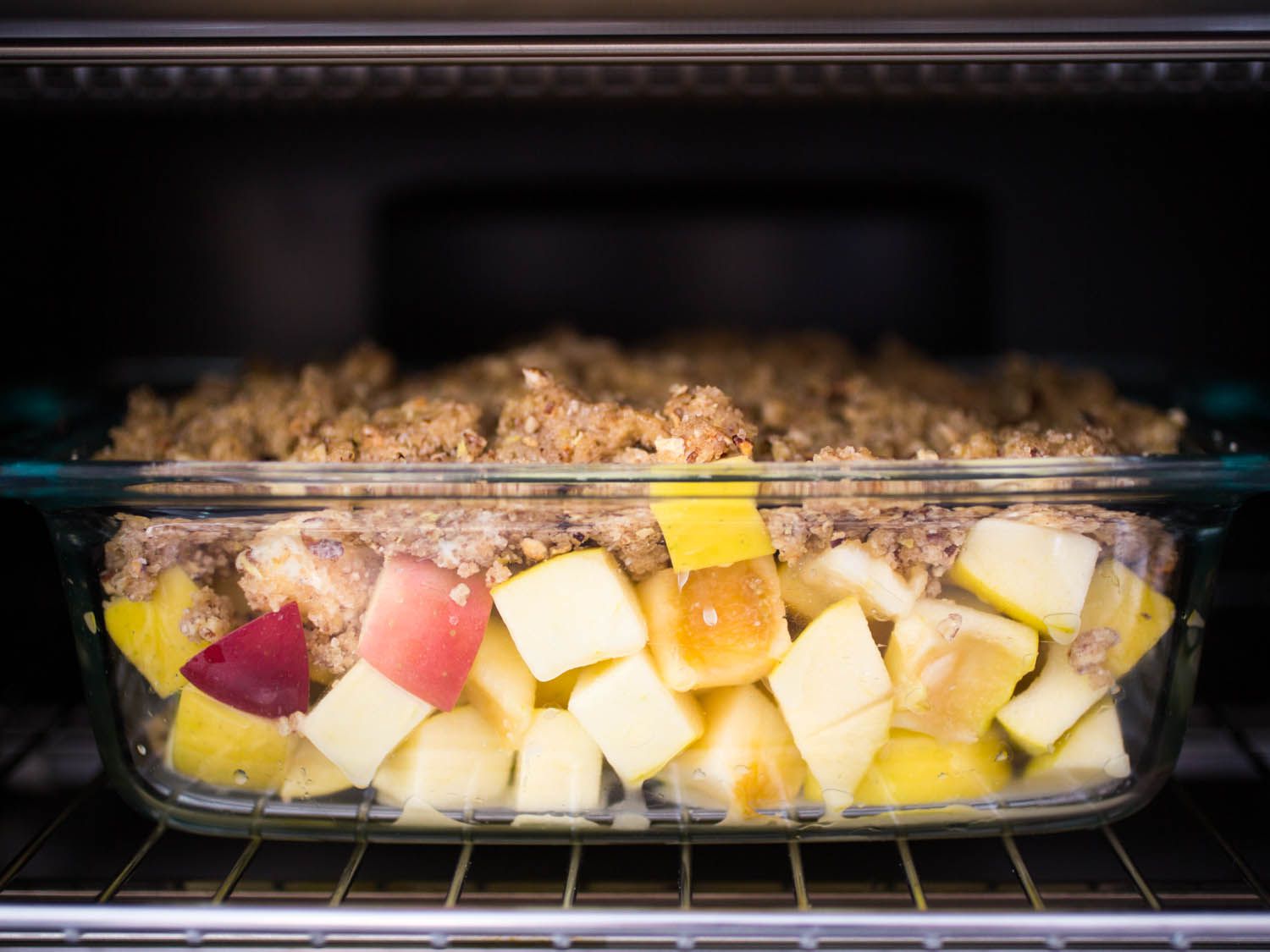 A side view of a glass baking dish of unbaked apple crisp, with chunks of red and yellow apples and a crumb topping, on an oven rack.