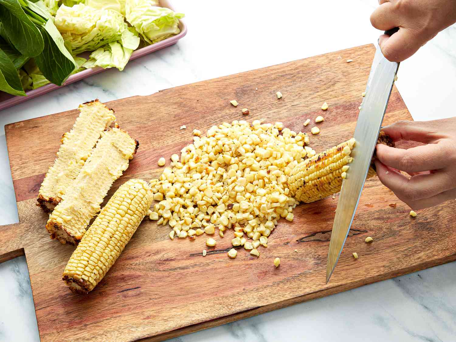 Slicing cooked corn off the cobs with a knife onto a wooden chopping board 