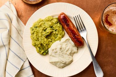 A plate with mashed peas, mashed potatoes, a sausage, and a fork resting on the edge