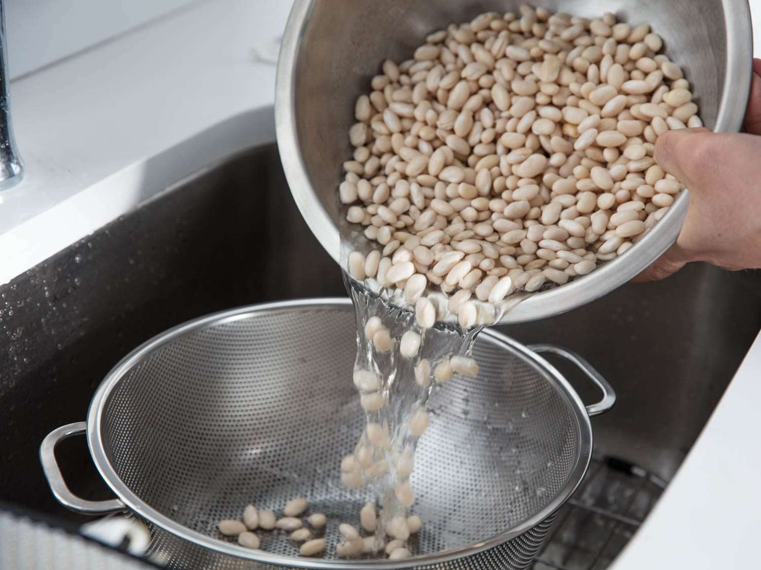 Pouring a large bowl of soaked white beans into a colander in a sink 