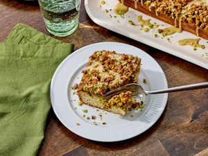 A slice of pistachio coffee cake on a plate with a fork a green napkin and glass of water nearby tray with more cake in the background