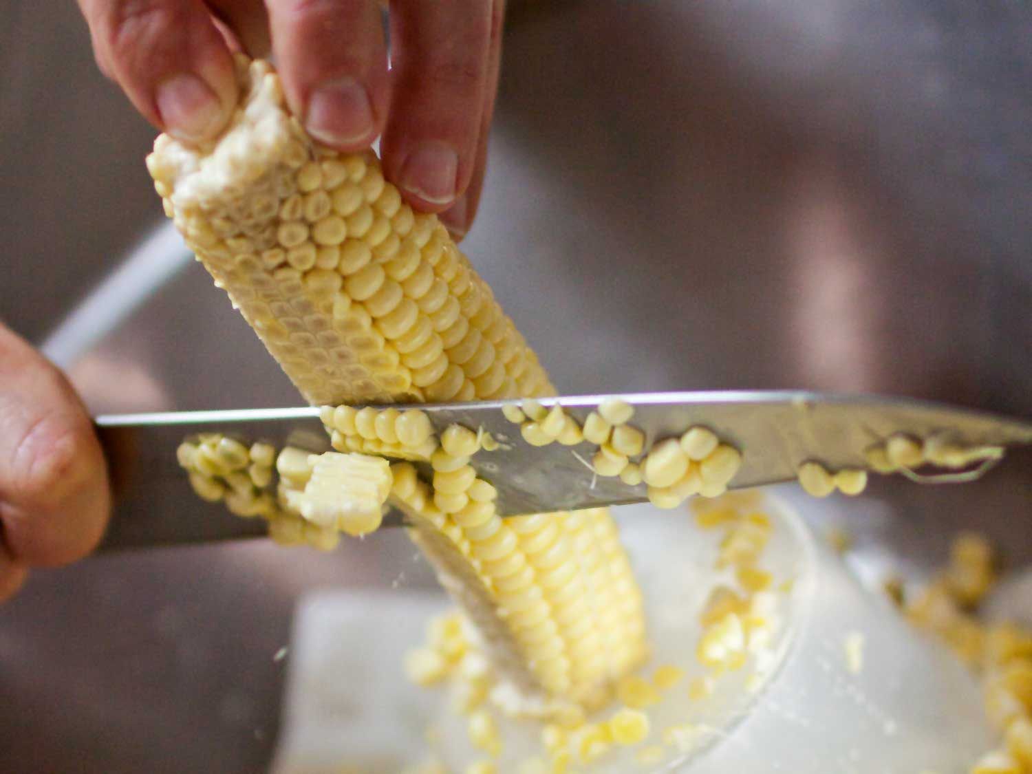 Corn kernels being cut off a cob with a chef's knife.