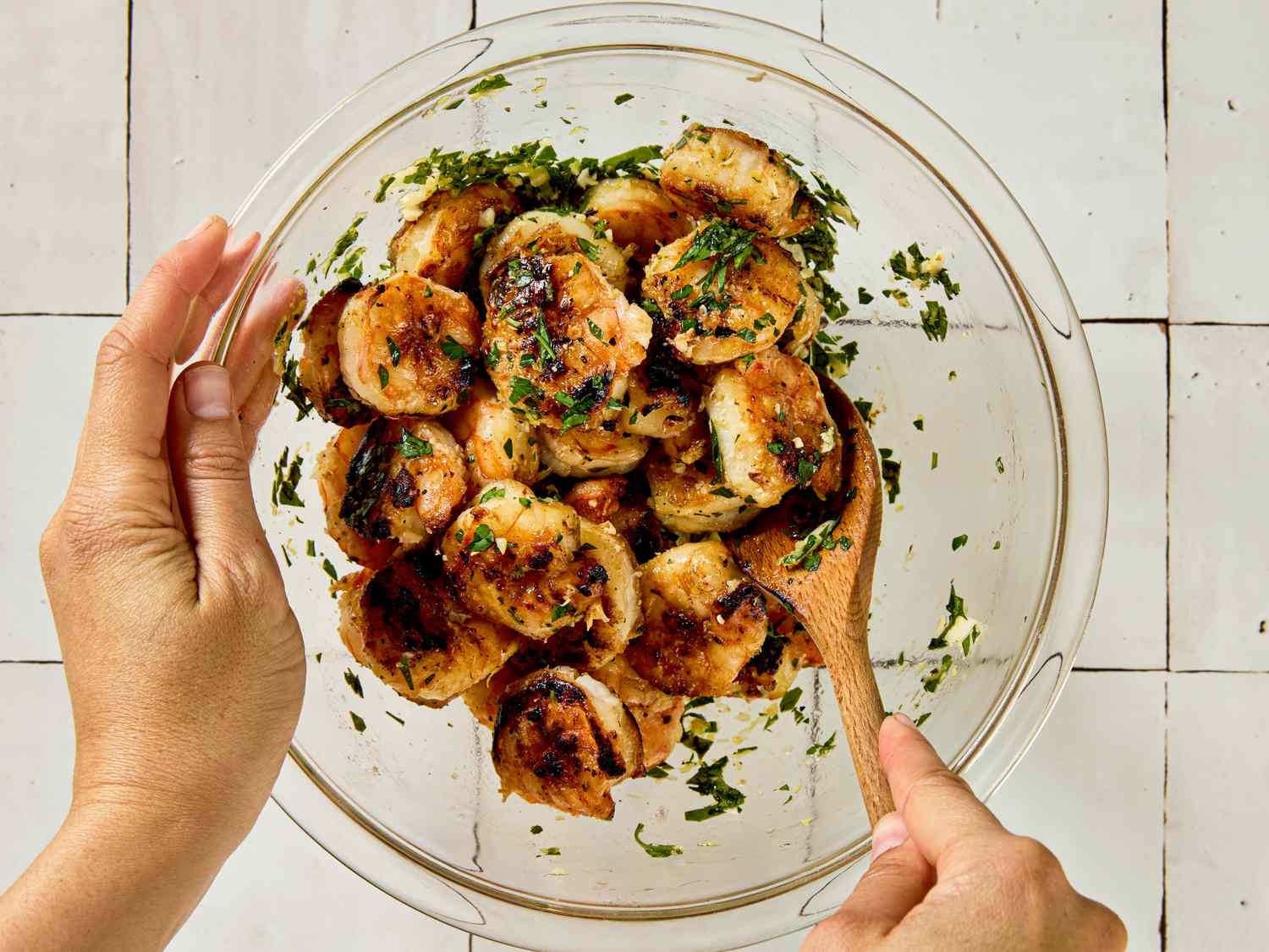 Hands mixing cooked shrimp with herbs in a glass bowl