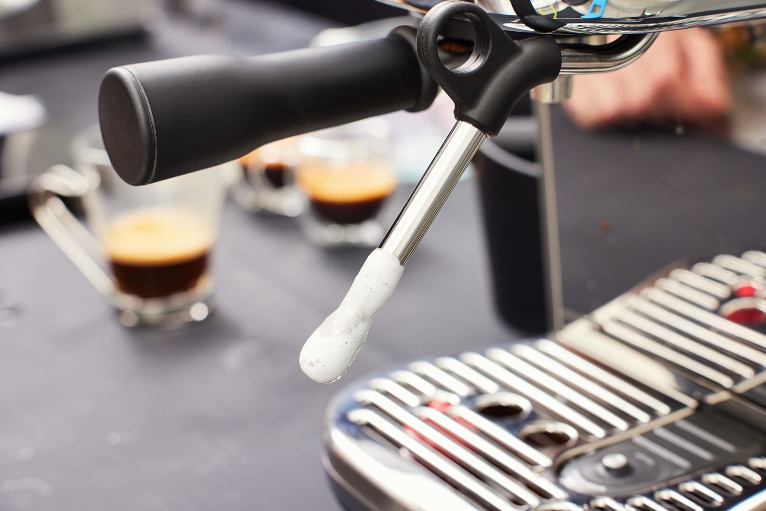 Closeup of Breville Bambino Plus Espresso Machine handle and steamer wand with two cups of coffee in the background