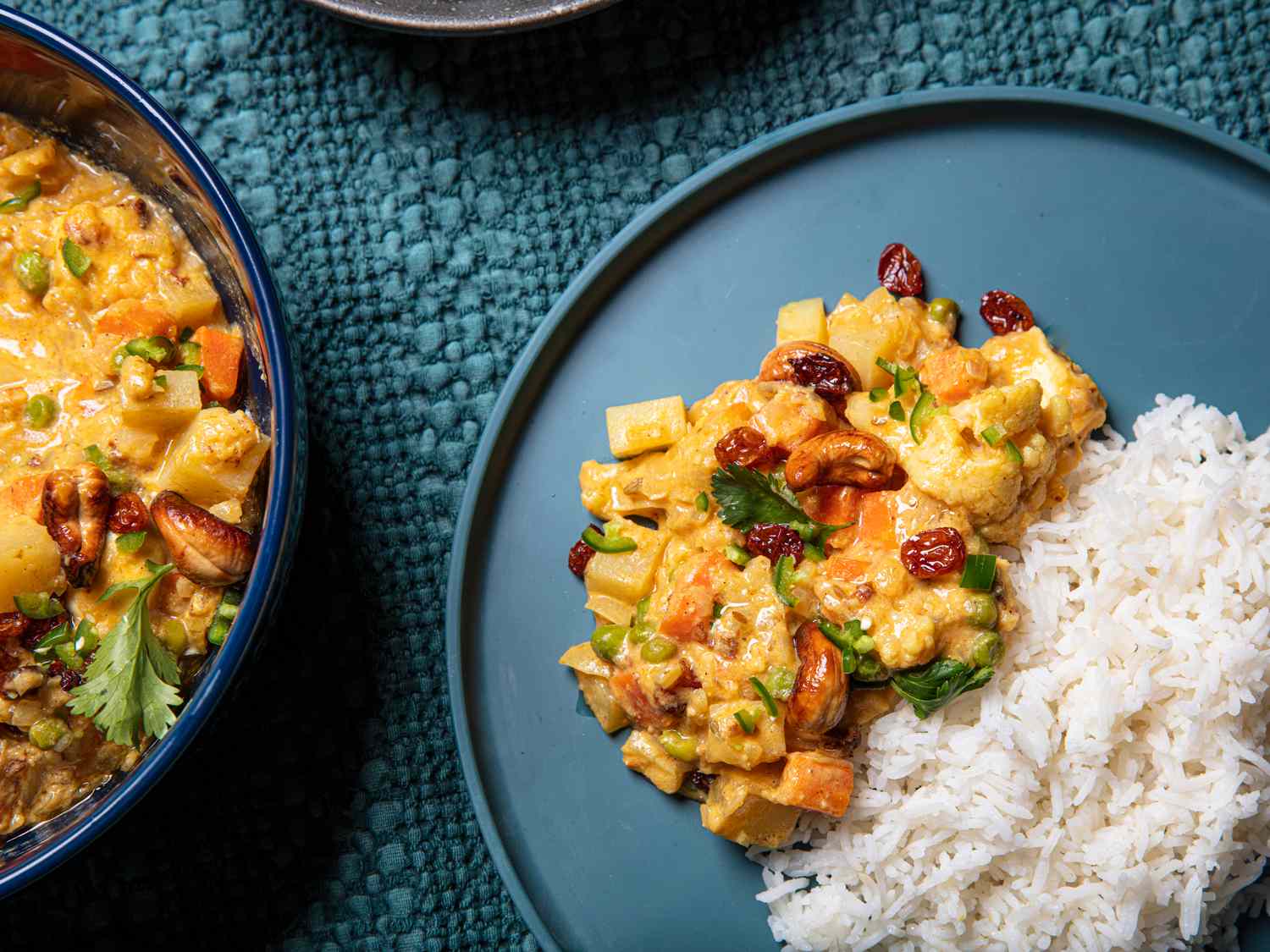Overhead view of a portion of vegetable qorma served next to white rice on a blue plate, on a textured blue fabric