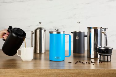 Five French press coffee makers on a wooden countertop. A person is pouring coffee from a French press into a mug.