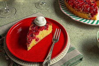 A slice of cranberry upsidedown cake on a red plate with a dollop of cream fork placed alongside the slice whole cake in background