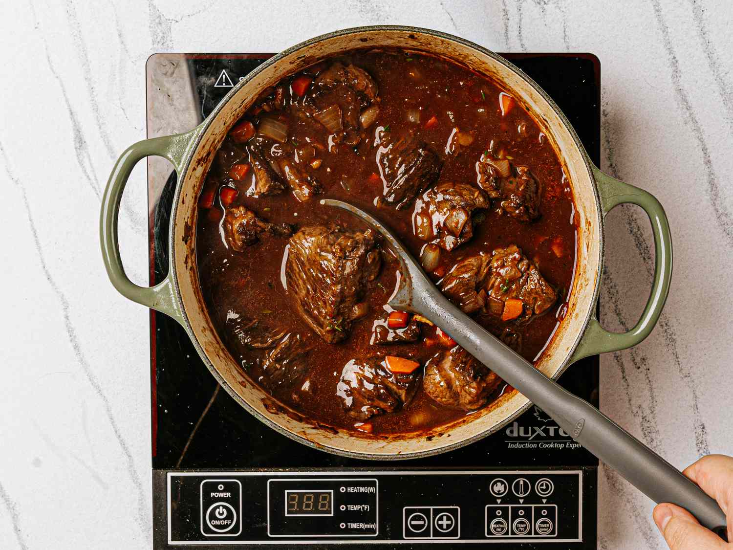 A pot of beef bourguignon cooking on an induction stove with a hand stirring using a ladle